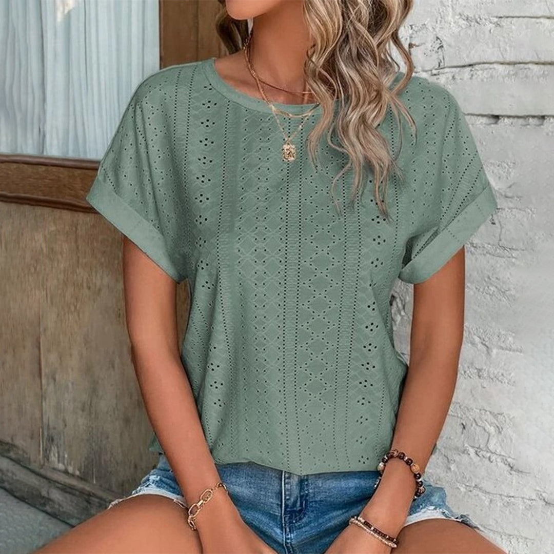 Vrouw draagt groene geperforeerde blouse met korte mouwen en denim shorts, zittend voor een witte muur. Trendy zomermode, casual dameskleding.