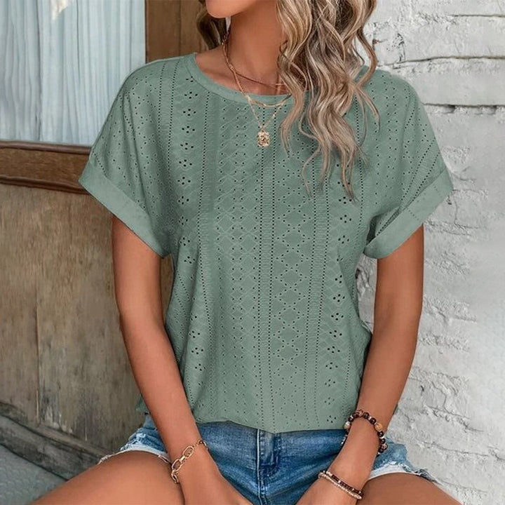Vrouw draagt groene geperforeerde blouse met korte mouwen en denim shorts, zittend voor een witte muur. Trendy zomermode, casual dameskleding.