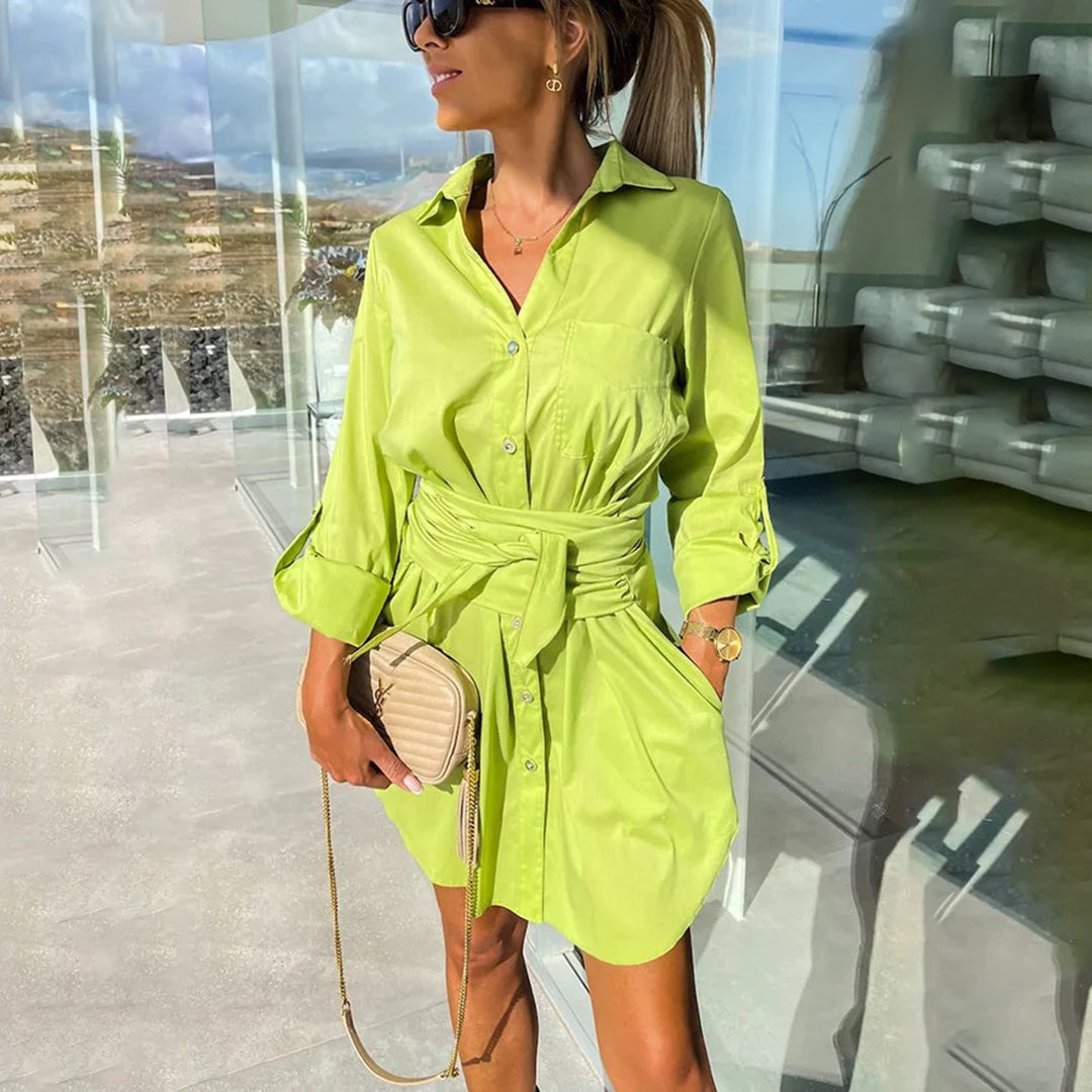 Vrouw in groene overhemdjurk met ceintuur, zonnebril en beige handtas, staand in moderne glazen ruimte met uitzicht op landschap.