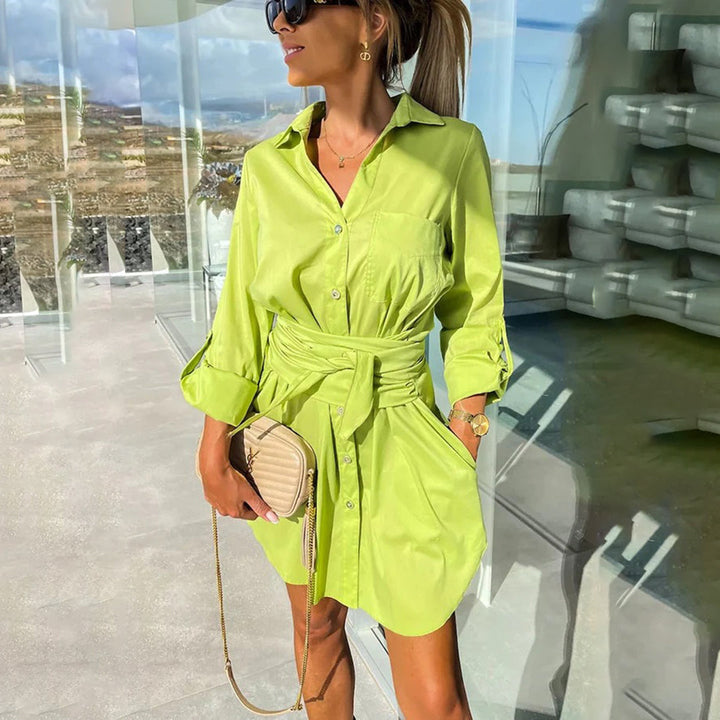 Vrouw in groene overhemdjurk met ceintuur, zonnebril en beige handtas, staand in moderne glazen ruimte met uitzicht op landschap.