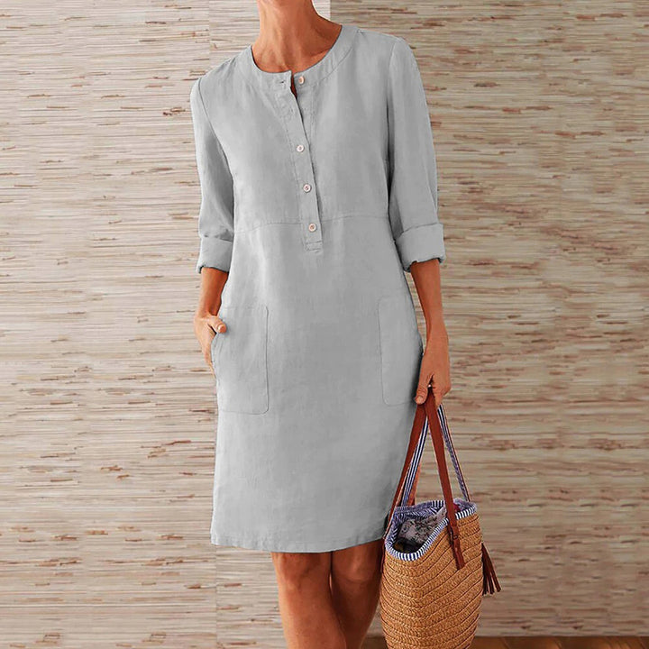 Grijze linnen jurk met knoopsluiting en opgerolde mouwen, gedragen door vrouw met rieten tas. Casual damesmode, comfortabele zomerjurk.