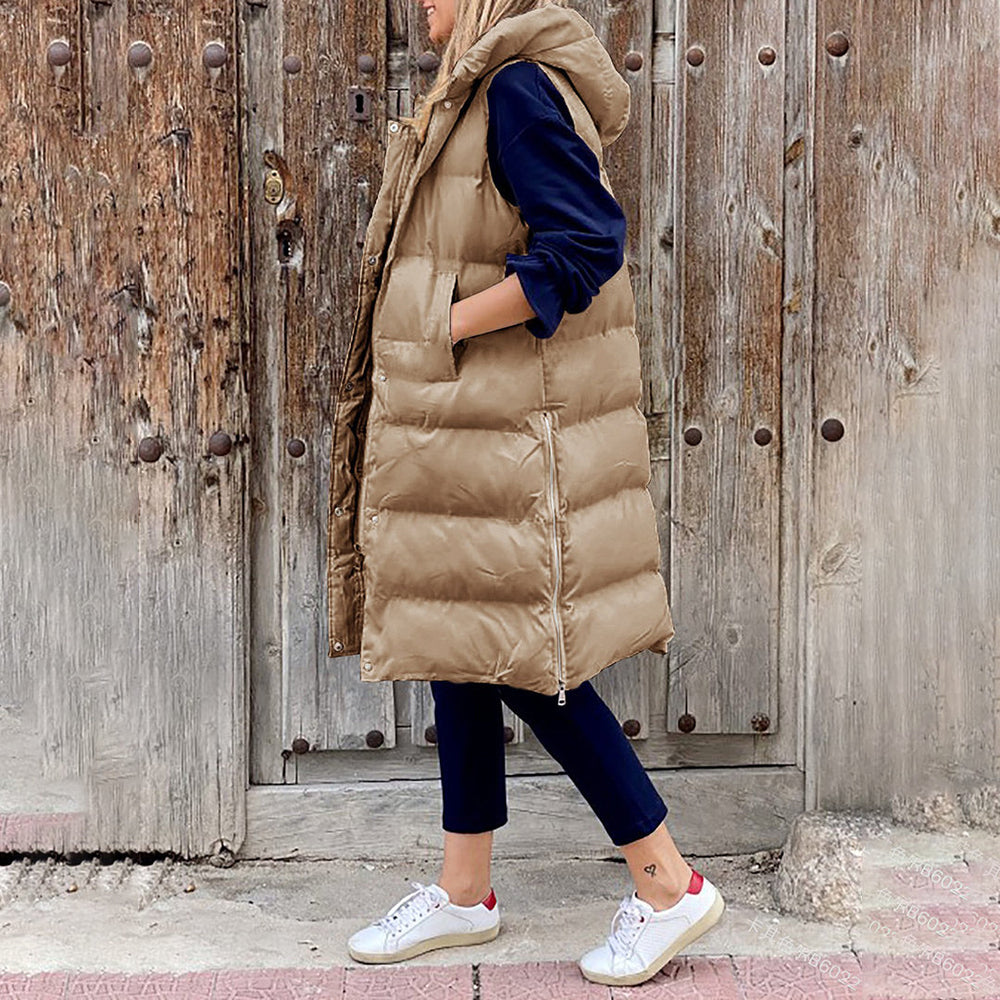 Vrouw in lange kaki gewatteerde bodywarmer, blauwe broek en witte sneakers, staand voor een rustieke houten deur. Wintermode, casual outfit.