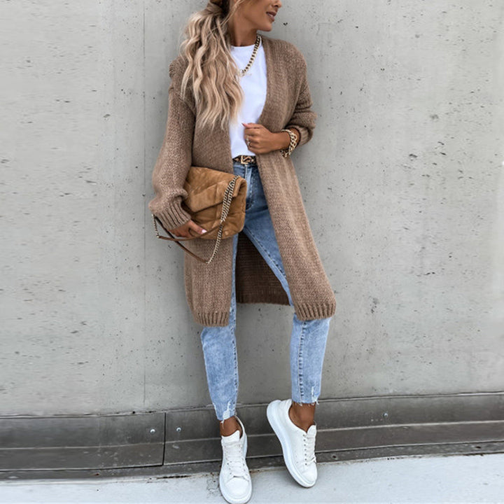 Vrouw in casual outfit met lange beige cardigan, witte sneakers en blauwe jeans, poseert tegen een betonnen muur. Modieuze herfstkledingstijl.