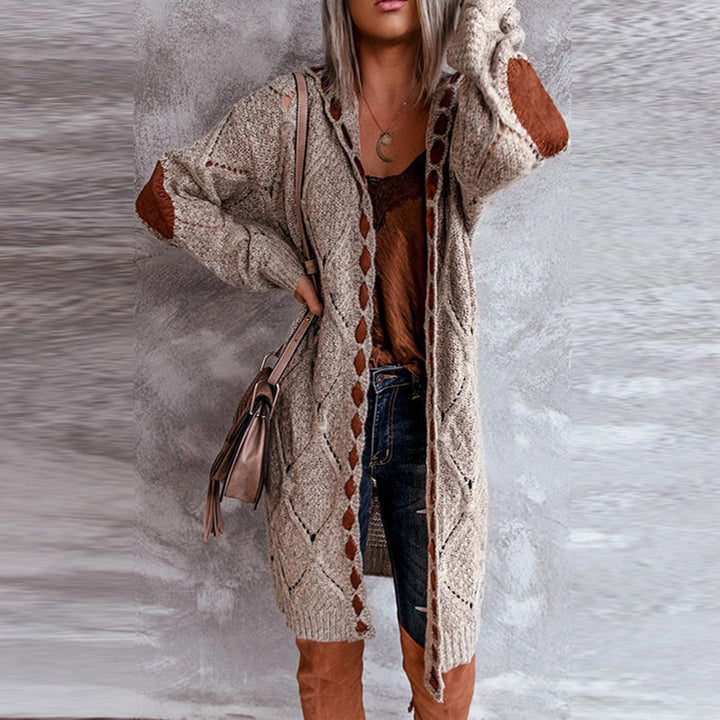 Vrouw in lange beige gebreide cardigan met bruine details, draagt jeans en bruine laarzen, poseert tegen grijze achtergrond. Modieuze herfstkleding.