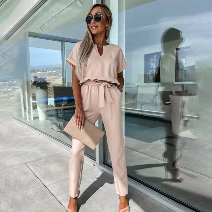 Vrouw in stijlvolle khaki jumpsuit met korte mouwen en strikceintuur, draagt zonnebril en clutch, poseert voor moderne glazen gevel.