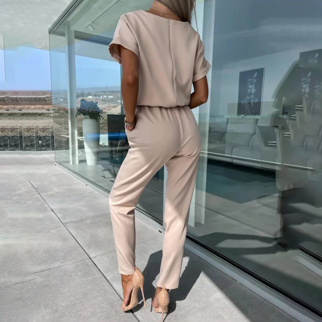Vrouw in stijlvolle kaki jumpsuit met korte mouwen, staand op modern terras met glazen wand. Modieuze dameskleding, zomeroutfit, elegant design.
