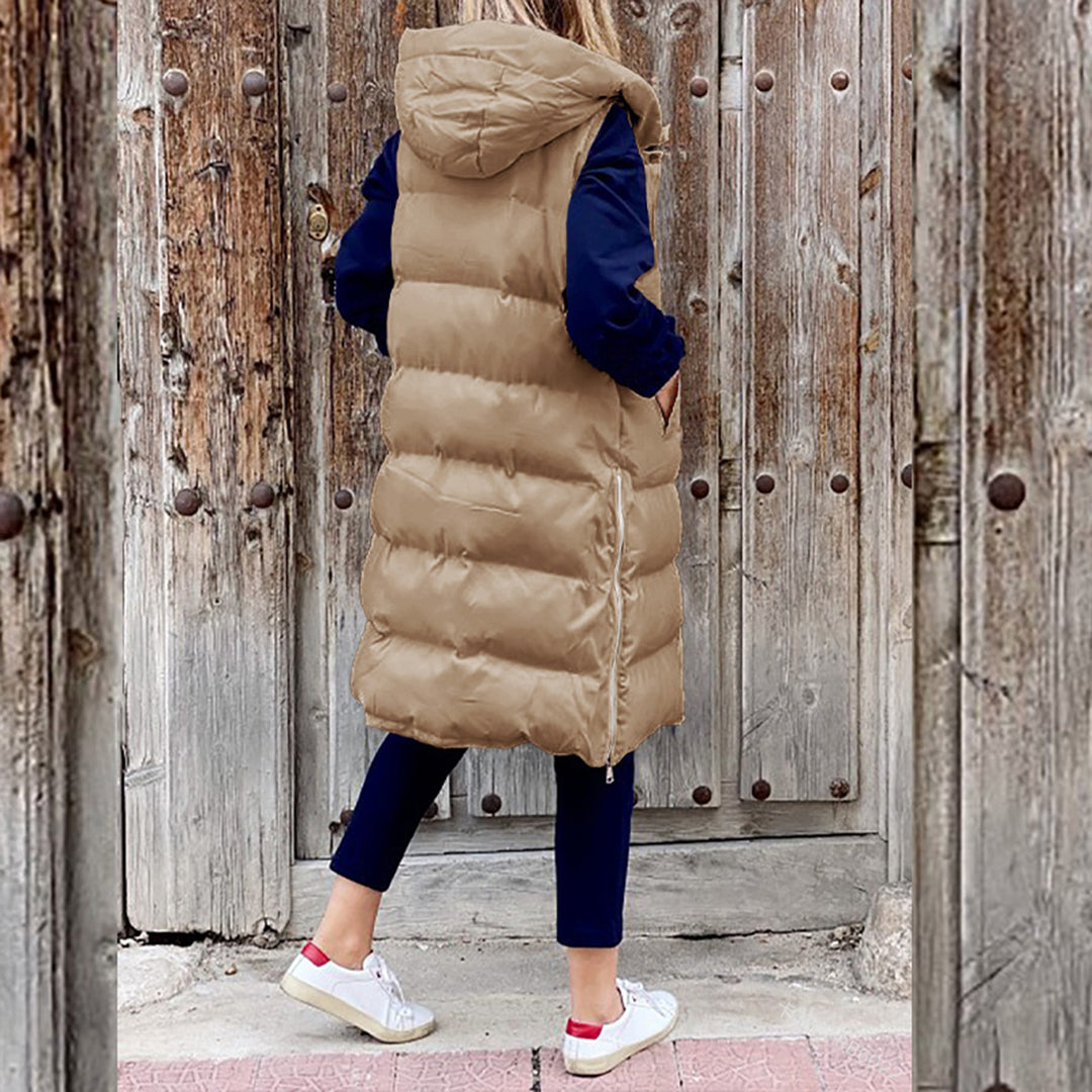 Vrouw in lange, kaki gewatteerde bodywarmer met capuchon, blauwe mouwen en witte sneakers, staand voor rustieke houten deur. Wintermode, casual stijl.