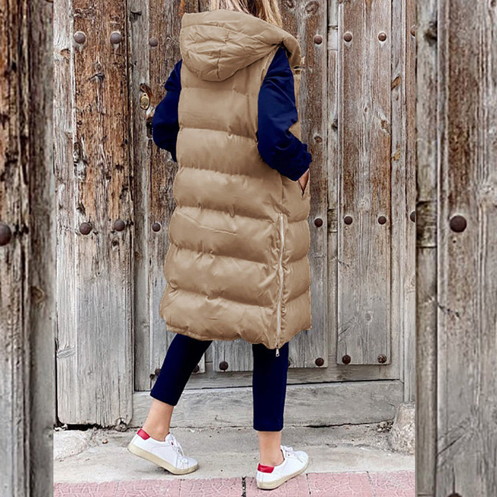 Vrouw in lange, kaki gewatteerde bodywarmer met capuchon, blauwe mouwen en witte sneakers, staand voor rustieke houten deur. Wintermode, casual stijl.
