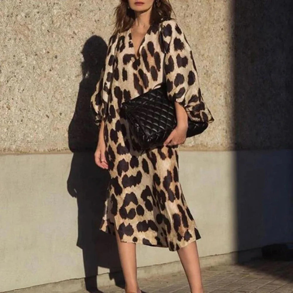 Vrouw in luipaardprint jurk met zwarte gewatteerde tas, staand tegen een betonnen muur. Modieuze dameskleding, trendy zomeroutfit.