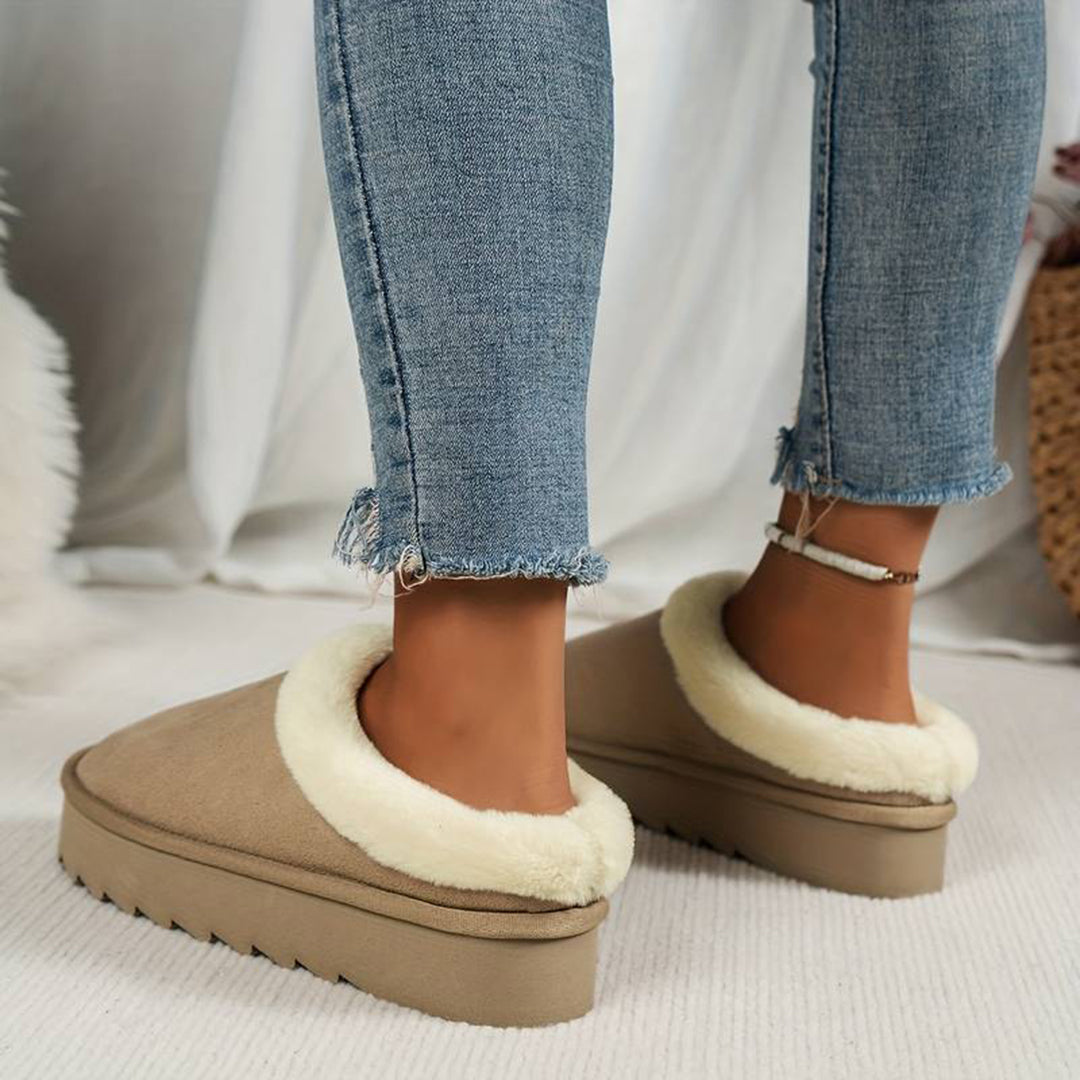 Vrouw draagt beige pantoffels met witte voering en dikke zool, gecombineerd met blauwe jeans. Comfortabele wintermode, warme damespantoffels.