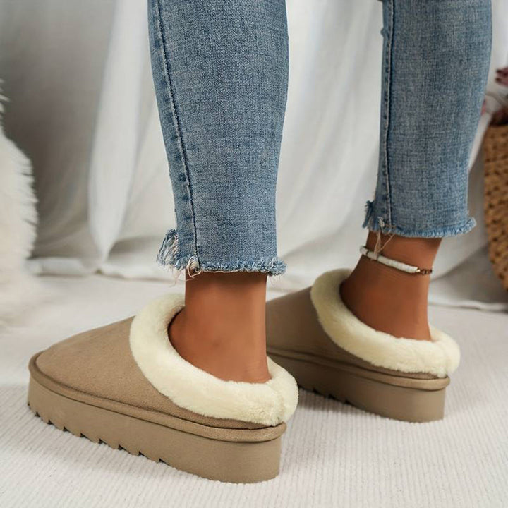 Vrouw draagt beige pantoffels met witte voering en dikke zool, gecombineerd met blauwe jeans. Comfortabele wintermode, warme damespantoffels.