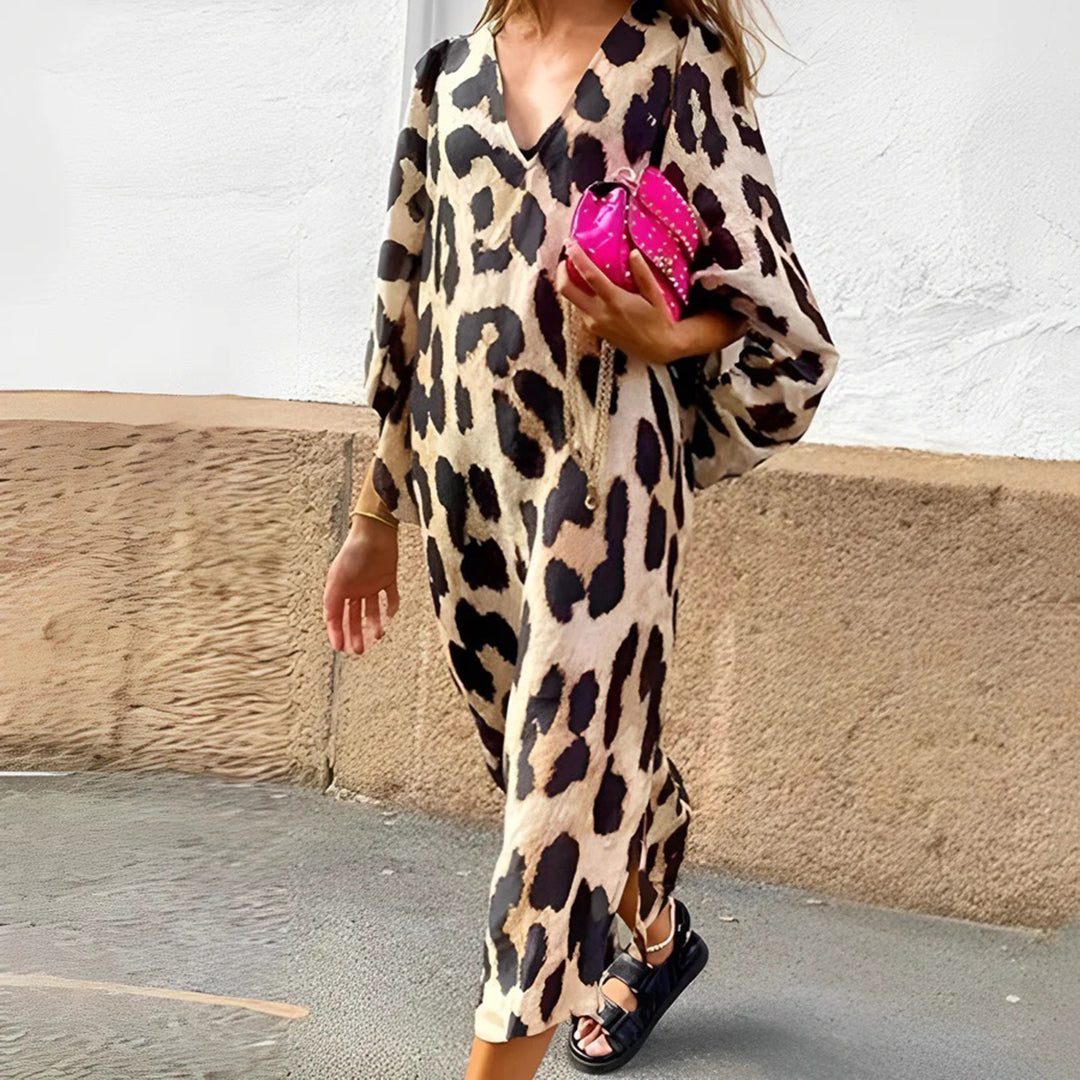 Vrouw in luipaardprint jurk met roze handtas en zwarte sandalen, loopt langs witte muur. Modieuze dameskleding, trendy accessoires, casual stijl.