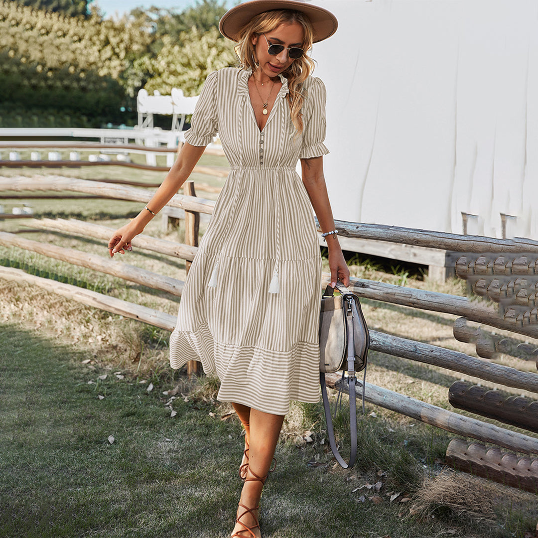 Vrouw in gestreepte kaki zomerjurk met hoed en zonnebril, wandelend langs houten hek. Perfecte boho-chic outfit voor zomerse dagen.