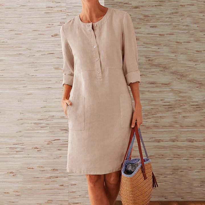 Vrouw in beige linnen jurk met knoopsluiting en opgerolde mouwen, houdt een strooien tas vast. Perfect voor casual zomeroutfits.