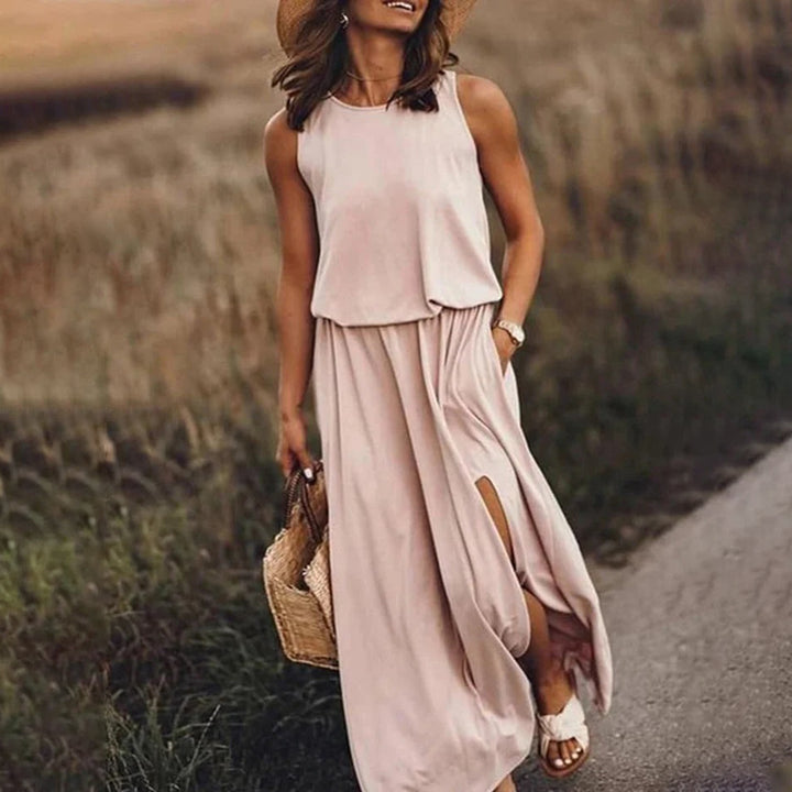 Vrouw in een elegante, mouwloze beige maxi-jurk met hoge split, draagt een strohoed en rieten tas, wandelend op een landelijk pad.