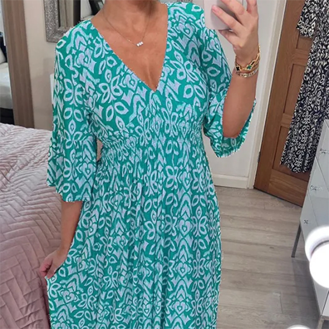 Vrouw in turquoise zomerjurk met bloemenpatroon, V-hals en driekwart mouwen, maakt selfie in slaapkamer. Trendy damesmode, casual stijl.