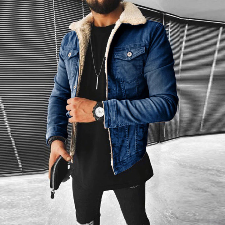 Man in blauwe spijkerjas met schapenvachtkraag, zwarte trui en horloge, houdt een zwarte portemonnee vast. Moderne herenkleding, casual stijl.