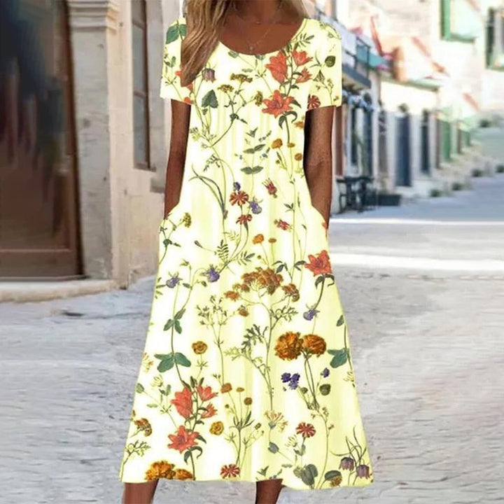 Vrouw in lichtgele zomerjurk met bloemenprint, korte mouwen en zakken, staand op een zonnige straat. Perfect voor casual zomermode.