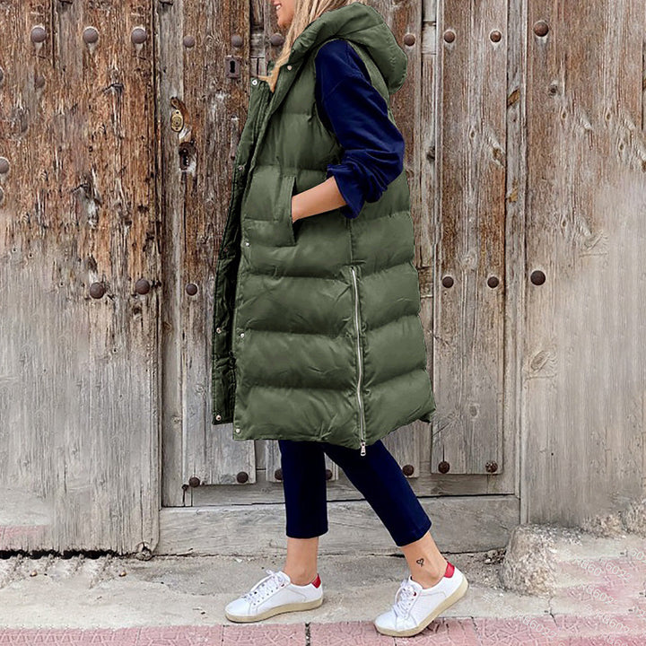Vrouw in groene gewatteerde bodywarmer met capuchon, donkerblauwe trui, zwarte broek en witte sneakers, staand voor houten deur.