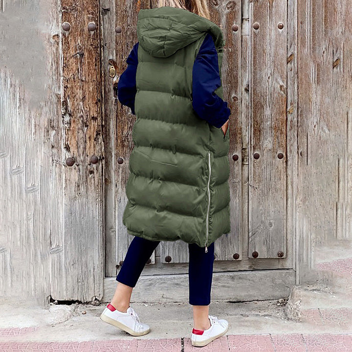 Vrouw in lange, groene gewatteerde bodywarmer met capuchon, voor houten deur. Modieuze winterkleding, casual stijl, comfortabele pasvorm.