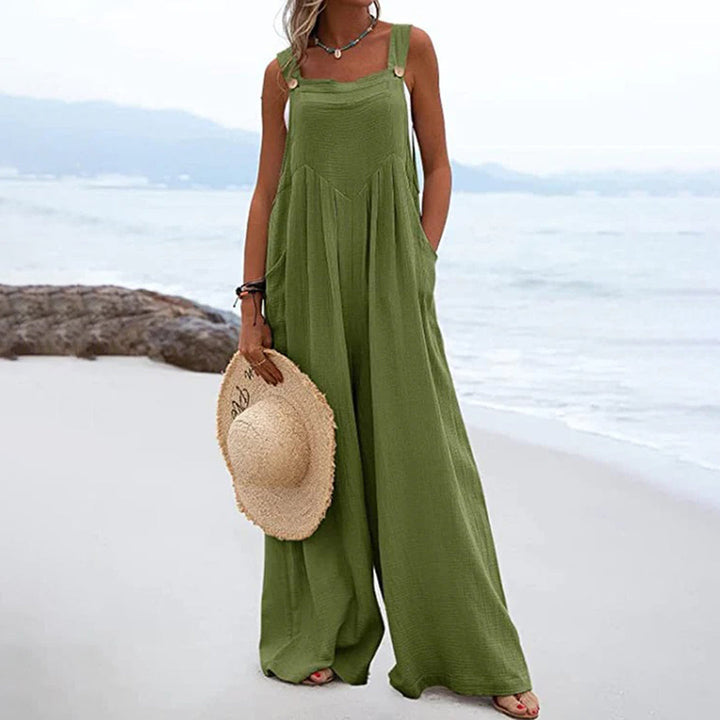 Vrouw in groene jumpsuit op strand, zomermode, casual strandkleding, wijde pijpen, trendy dameskleding, strandhoed, ontspannen stijl.
