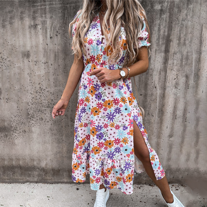 Vrouw in kleurrijke bloemenjurk met korte mouwen en split, poseert tegen betonnen muur. Trendy zomeroutfit, casual stijl, witte sneakers.