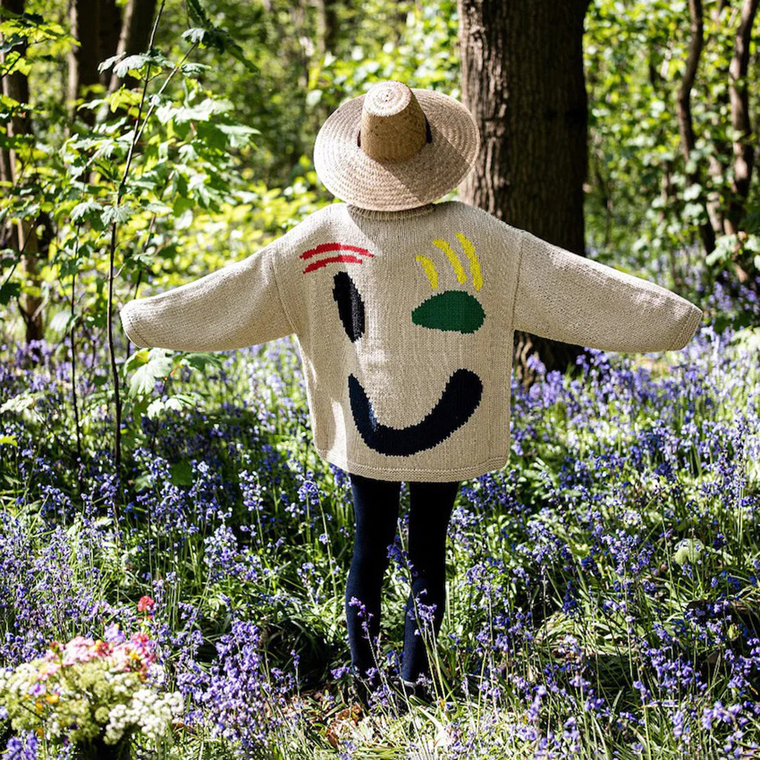 Kind in bos met strohoed en kleurrijke trui met gezichtsmotief, omringd door paarse bloemen en groene bomen, lente natuurwandeling.