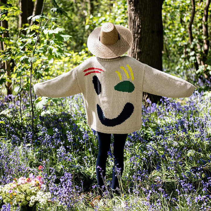 Kind in bos met strohoed en kleurrijke trui met gezichtsmotief, omringd door paarse bloemen en groene bomen, lente natuurwandeling.