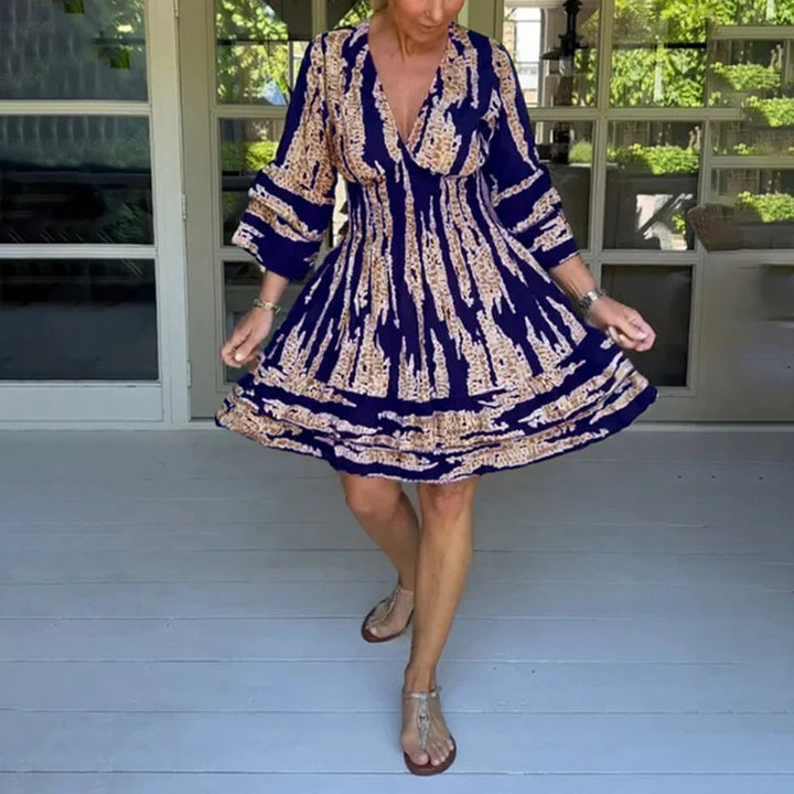 Vrouw draagt een marineblauwe zomerjurk met beige patroon, driekwart mouwen en V-hals, op een veranda. Perfect voor casual zomermode.