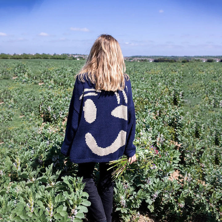 Vrouw in veld met donkerblauwe trui met grote smiley, omringd door groene planten. Trendy mode, casual kleding, buitenstijl.