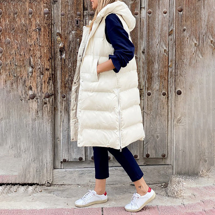 Vrouw draagt lange, off-white gewatteerde bodywarmer met capuchon, gecombineerd met donkerblauwe kleding en witte sneakers, voor houten deur.