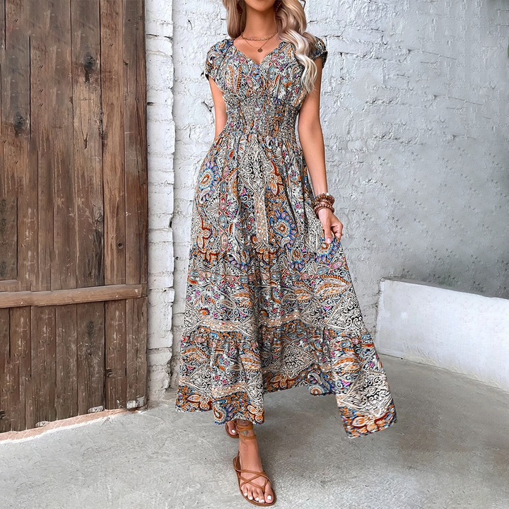 Vrouw in kleurrijke bohemian maxi-jurk met paisley-patroon, staand tegen een rustieke houten deur en witte stenen muur, zomermode.