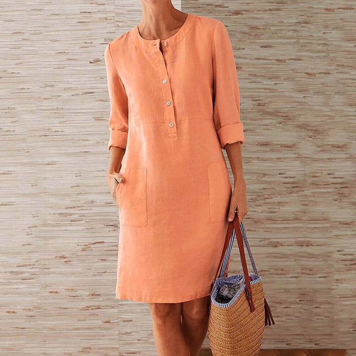 Oranje linnen jurk met knoopsluiting en opgerolde mouwen, gedragen door een vrouw met een rieten strandtas. Perfect voor zomerse casual outfits.