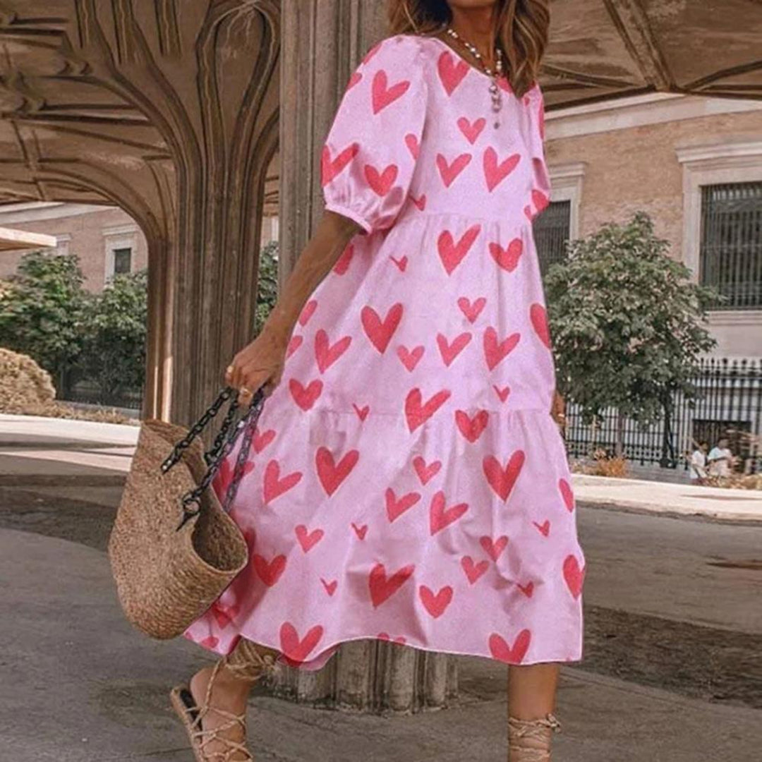 Roze zomerjurk met rode hartjesprint, gedragen door vrouw met rieten tas. Perfect voor casual zomermode en stranduitjes.