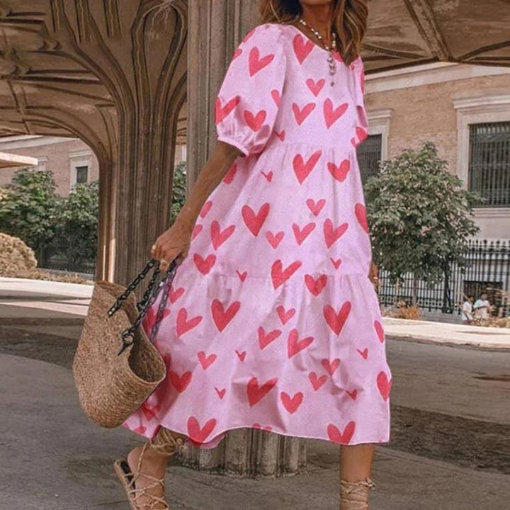 Roze zomerjurk met rode hartjesprint, gedragen door vrouw met rieten tas. Perfect voor casual zomermode en stranduitjes.