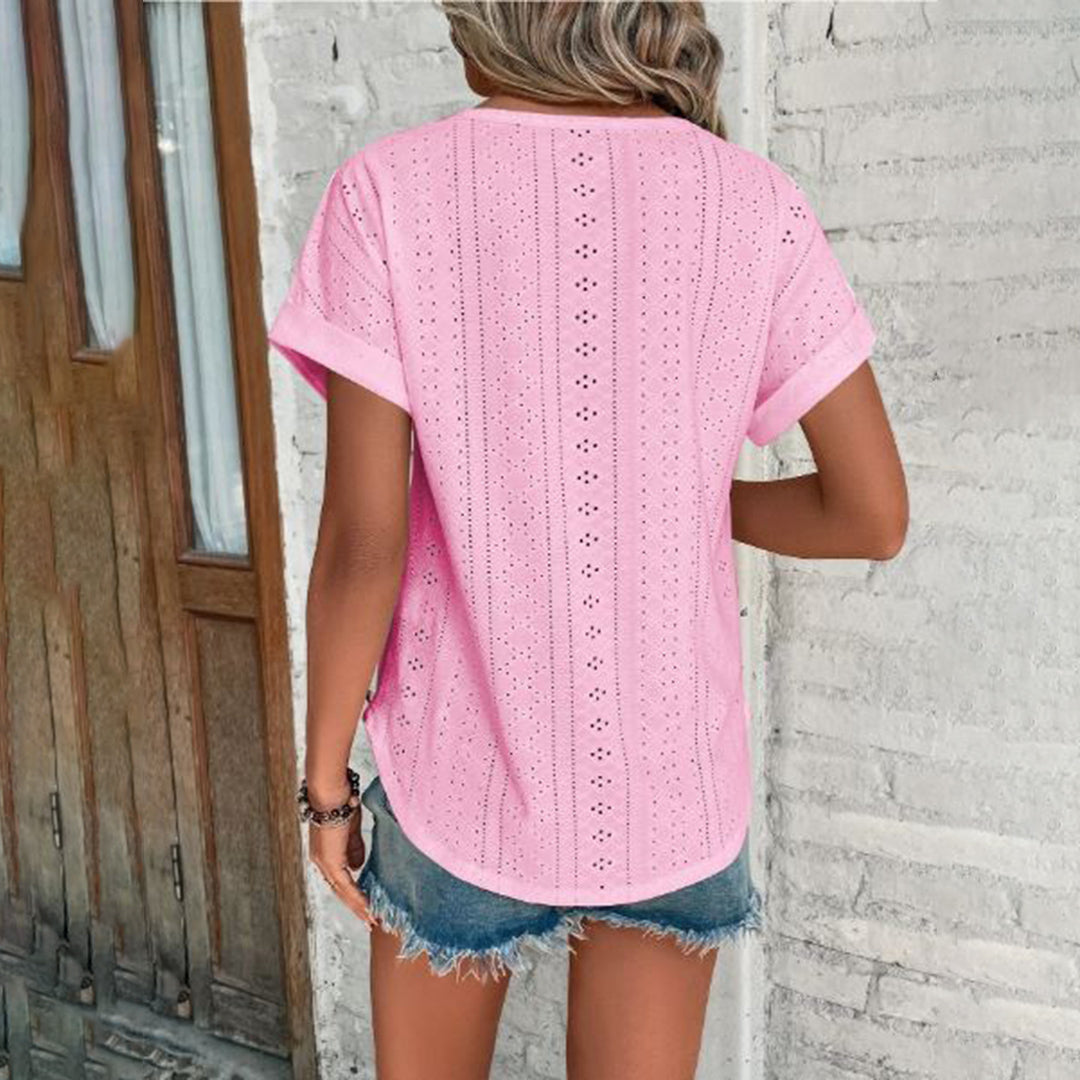 Vrouw in roze geperforeerde blouse en denim shorts, staand voor een witte bakstenen muur. Zomermode, casual outfit, trendy dameskleding.