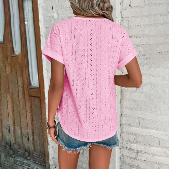 Vrouw in roze geperforeerde blouse en denim shorts, staand voor een witte bakstenen muur. Zomermode, casual outfit, trendy dameskleding.