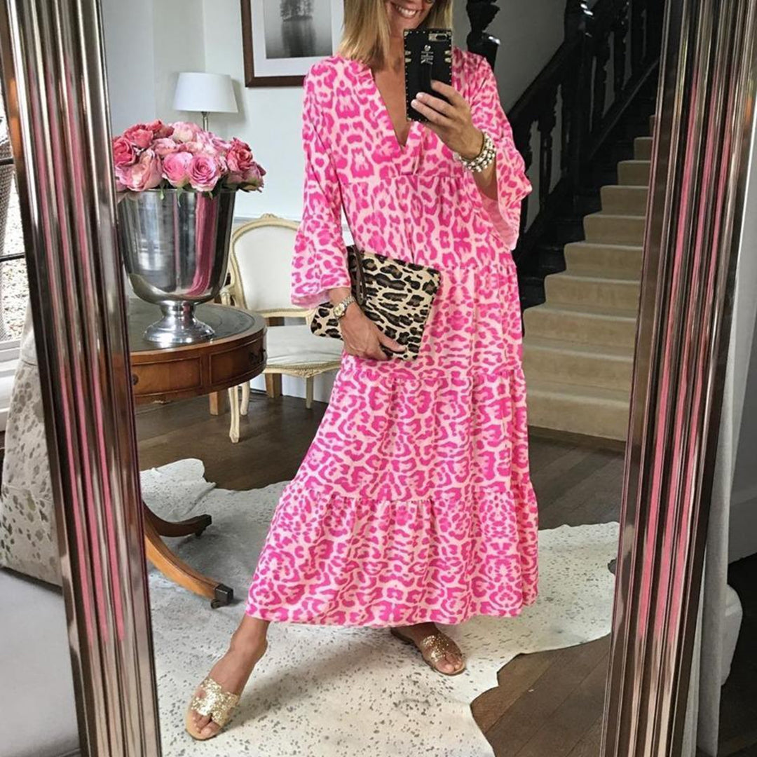 Vrouw in roze luipaardprint jurk poseert voor spiegel, met luipaardprint clutch en gouden sandalen. Luxe interieur met bloemen op achtergrond.