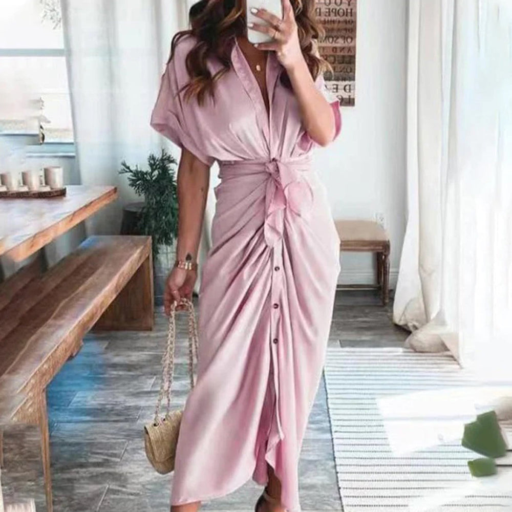 Vrouw in roze maxi-jurk met knopen en strikdetail, staand in moderne woonkamer. Trendy damesmode, zomerjurk, casual elegantie.