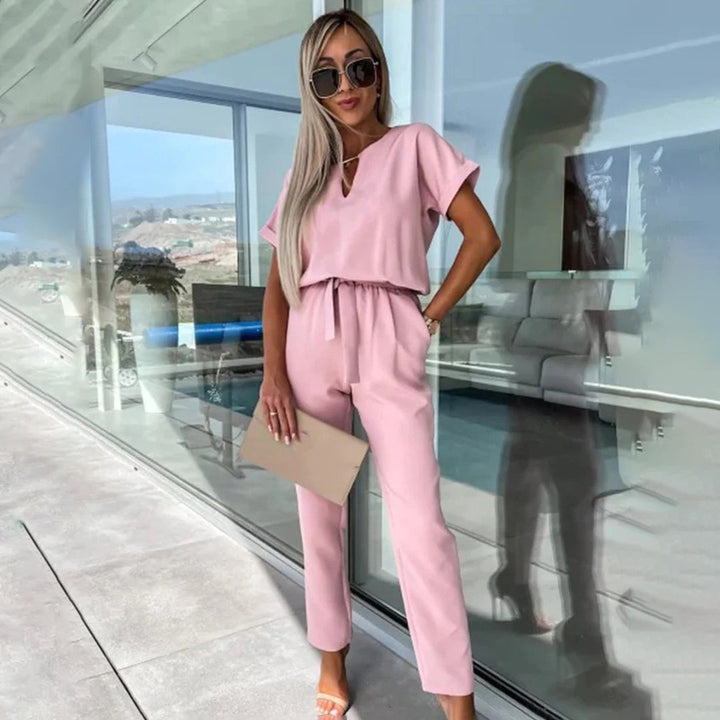 Vrouw in roze jumpsuit met zonnebril en clutch, staand bij glazen wand. Modieuze dameskleding, zomeroutfit, casual chic stijl.