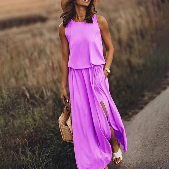 Vrouw in een lange, roze zomerjurk met hoge split, draagt een strohoed en rieten tas, wandelend op een landweg. Perfecte outfit voor zomermode.