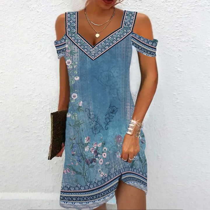 Vrouw in blauwe zomerjurk met bloemenprint en schouderbandjes, tegen witte achtergrond. Perfect voor casual zomeroutfits en strandmode.