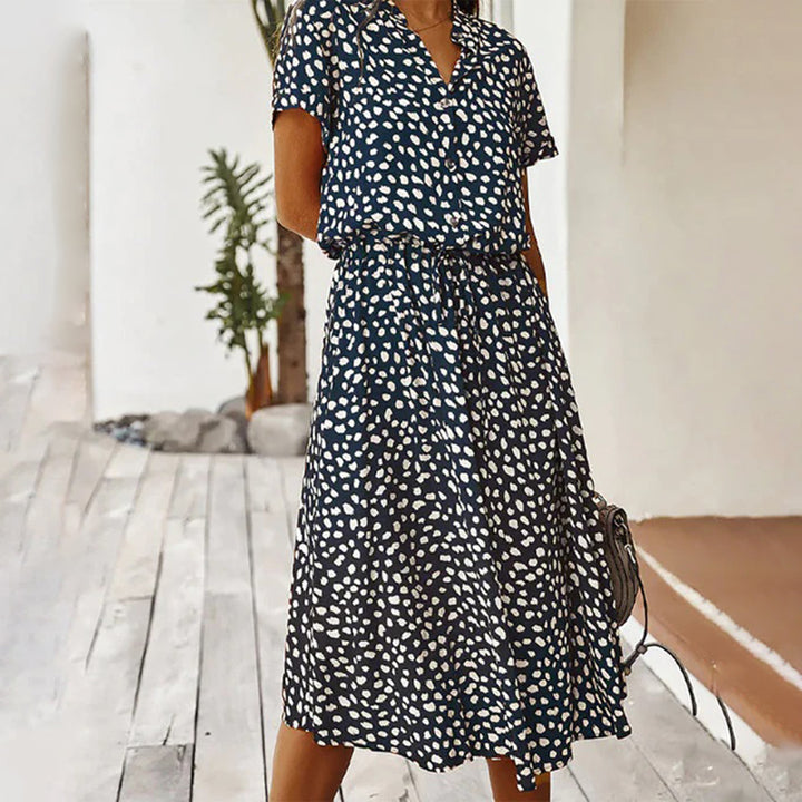 Vrouw in elegante, donkerblauwe jurk met witte stippen, korte mouwen en V-hals, staand op houten vloer. Perfect voor zomerse mode en casual stijl.