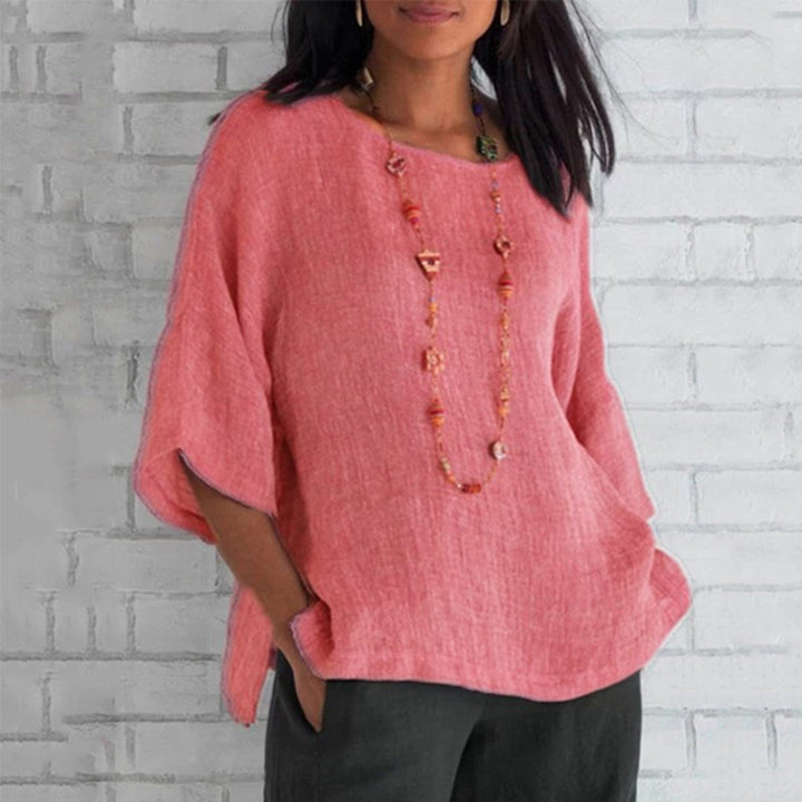 Vrouw draagt roze linnen blouse met korte mouwen en lange ketting, tegen witte bakstenen muur. Modieuze dameskleding, casual zomeroutfit.