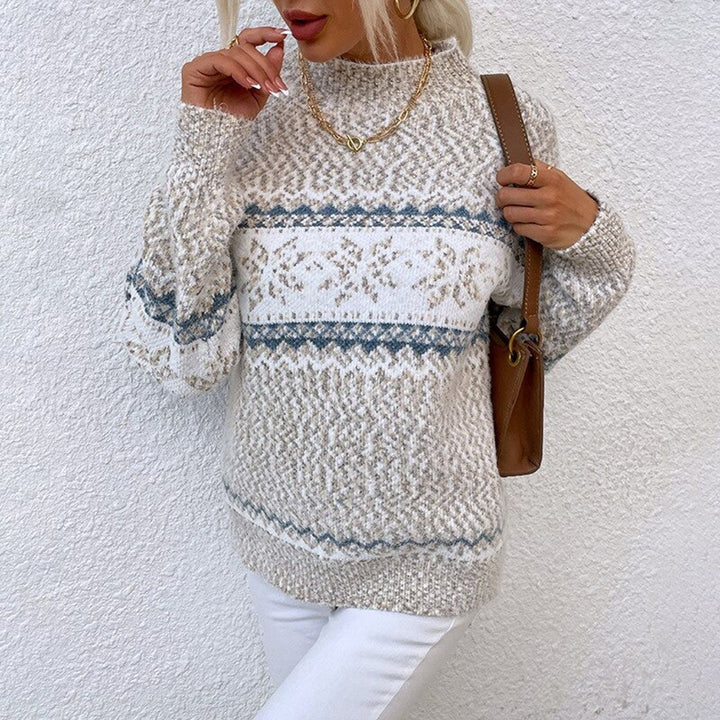 Vrouw draagt gebreide trui met Noors patroon, beige en blauw, gecombineerd met witte broek en bruine tas, tegen witte achtergrond.