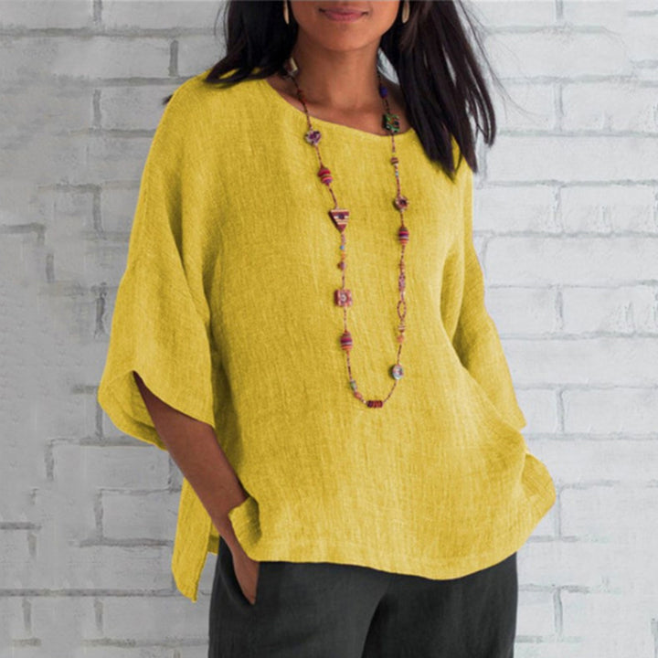 Vrouw draagt gele linnen tuniek met korte mouwen en kleurrijke ketting, tegen witte bakstenen muur. Modieuze dameskleding, casual stijl.