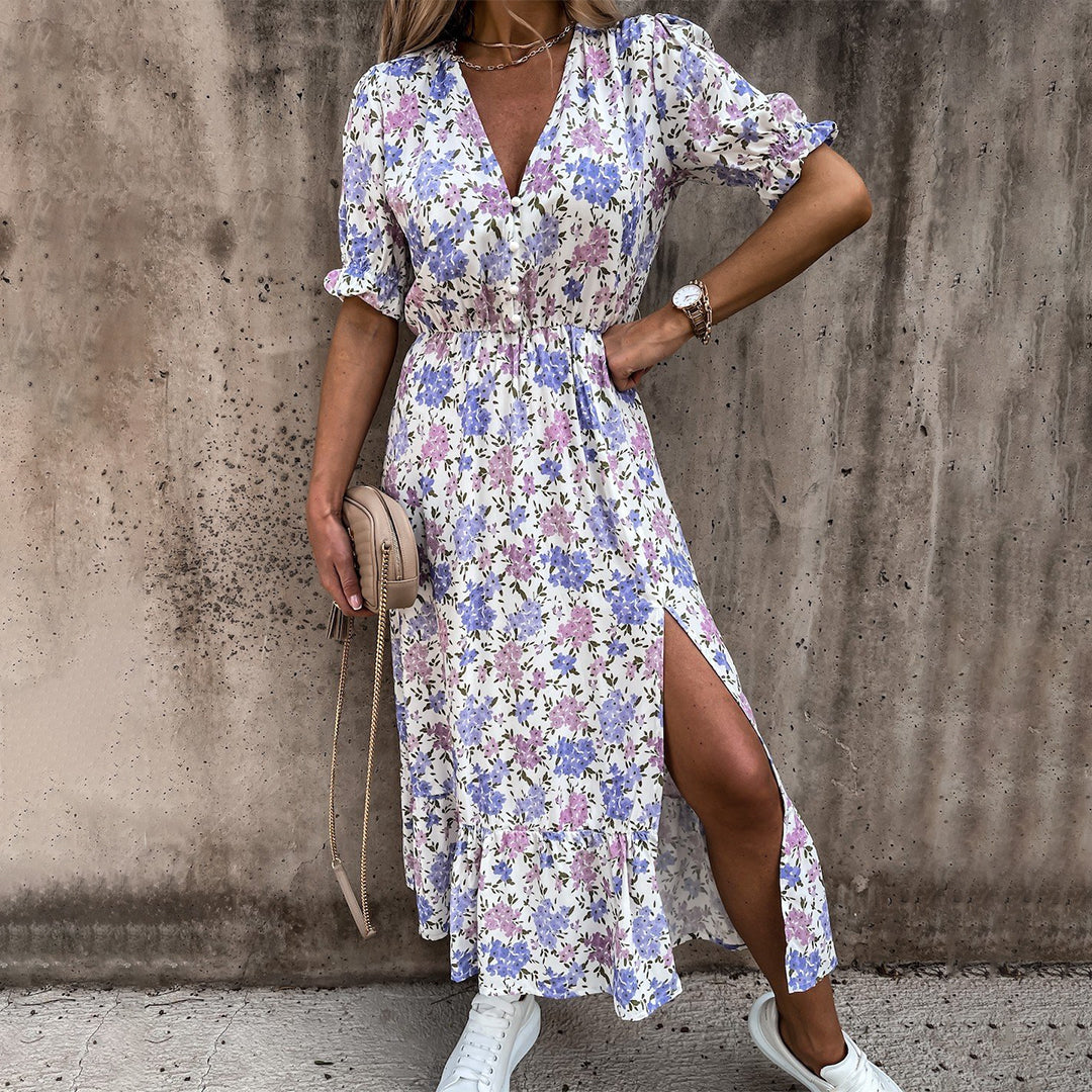 Vrouw in bloemenjurk met paarse en roze print, split aan de zijkant, witte sneakers en beige tas, tegen een betonnen muur. Zomermode, casual stijl.