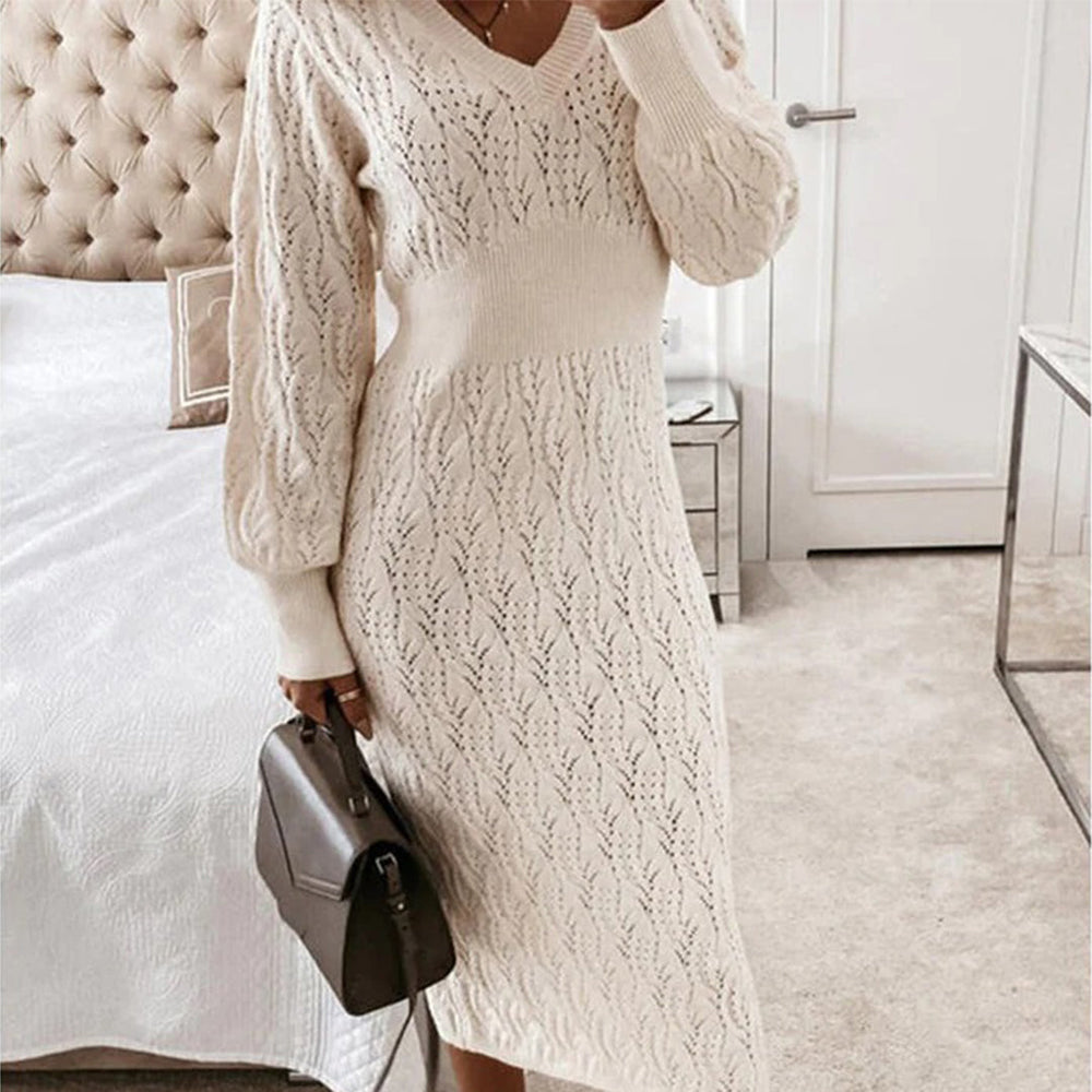 Vrouw in beige gebreide jurk met V-hals en lange mouwen, houdt een bruine handtas vast in een moderne slaapkamer. Elegante wintermode.