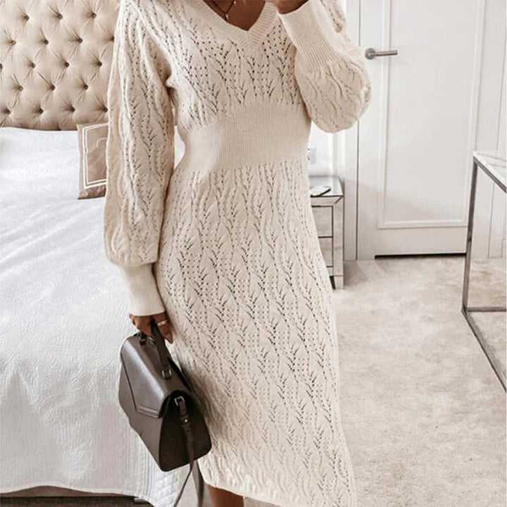 Vrouw in beige gebreide jurk met V-hals en lange mouwen, houdt een bruine handtas vast in een moderne slaapkamer. Elegante wintermode.