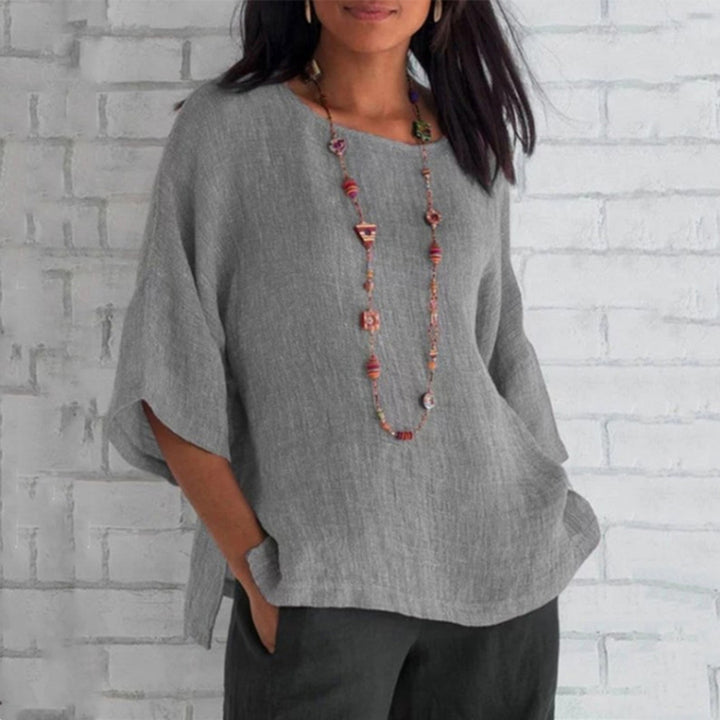Grijze linnen blouse voor dames, driekwart mouwen, casual stijl, gedragen met lange kleurrijke ketting, tegen witte bakstenen muur.